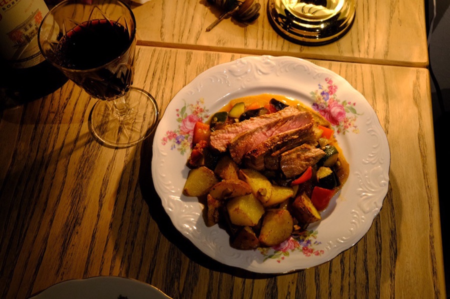 A home-cooked dinner of seared steak and roast vegetables on a floral plate, with a glass of red wine.